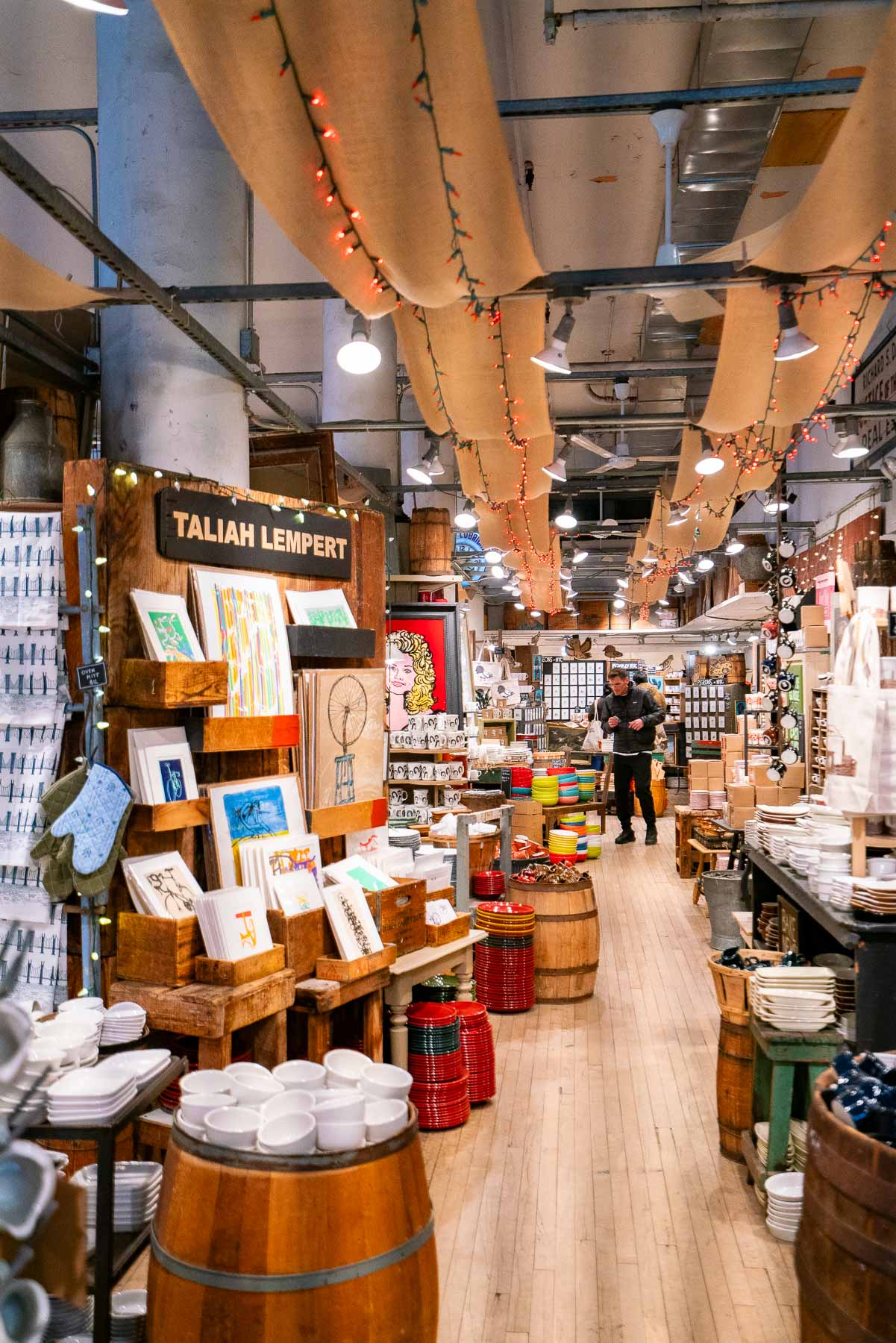 Man shopping for trinkets and artwork at Fishs Eddy. Walls full of artwork and vintage plates on display with string lights from the ceiling