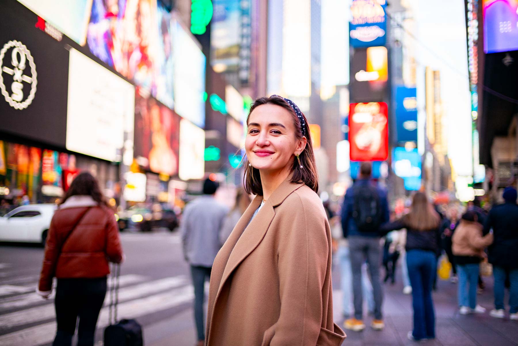 A woman in a camel coat stands in Times Square with illuminated billboards and pedestrians around her.