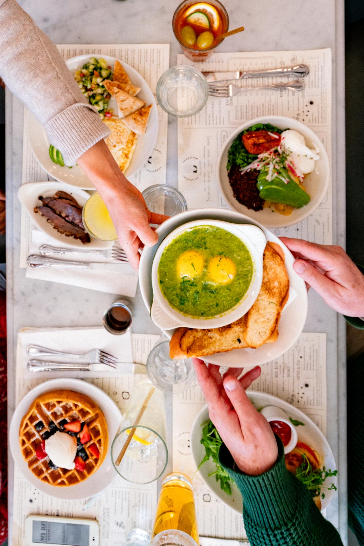 An overhead view of a brunch spread on a marble table with hands reaching for dishes.