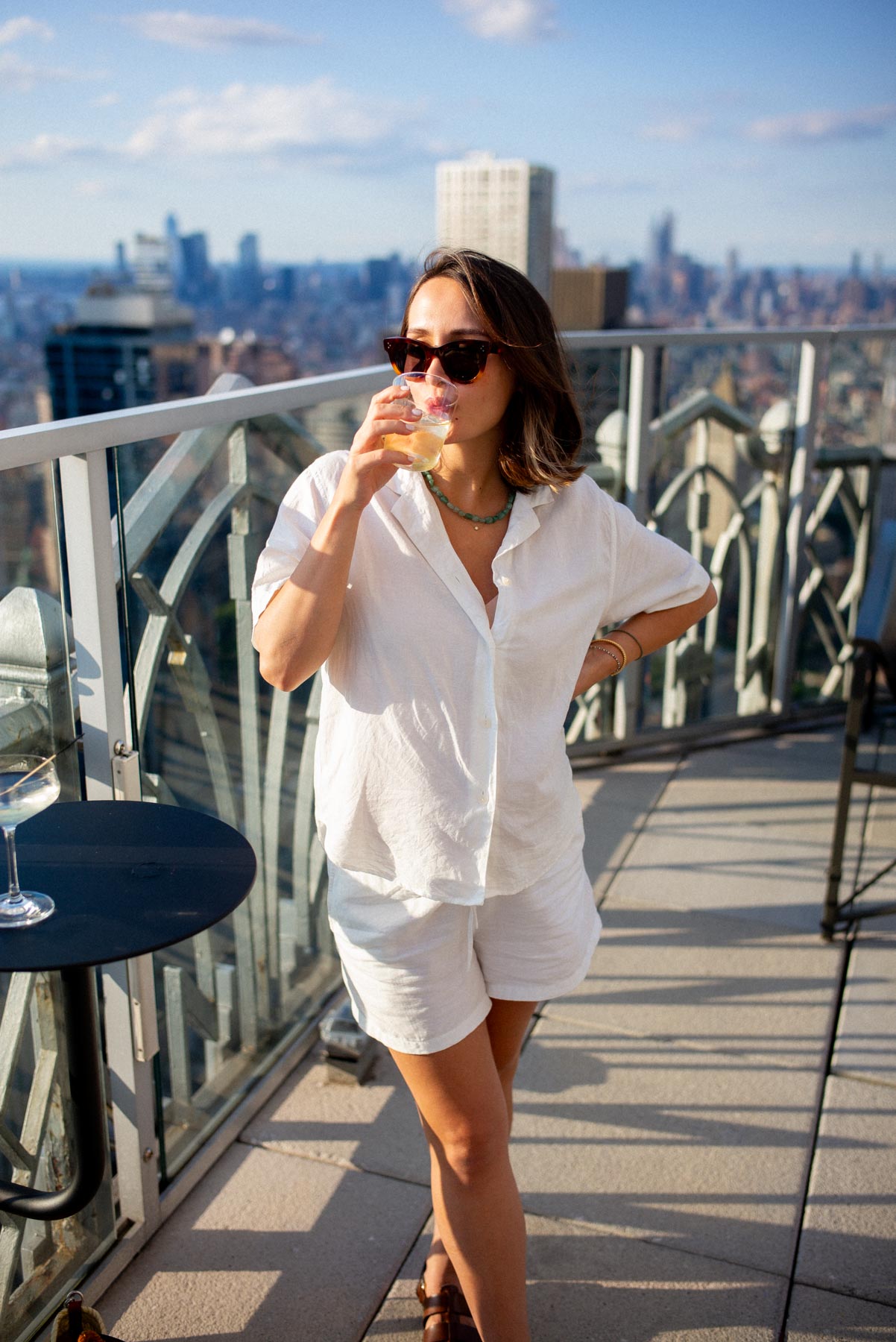 A woman drinks a cocktail on a rooftop bar overlooking the Manhattan skyline.