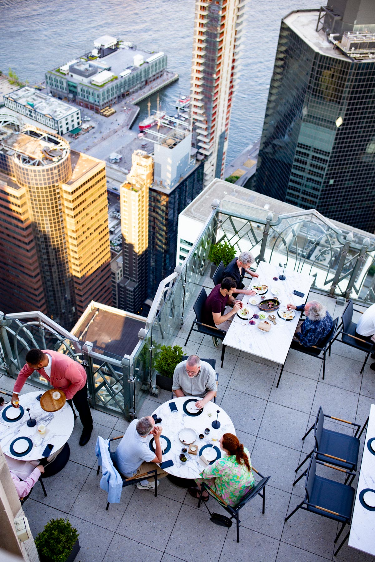 An aerial view shows diners seated at tables on a high-rise rooftop terrace overlooking the Financial District skyline and river.