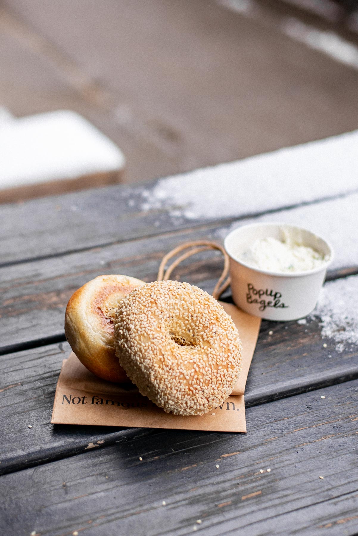 A sesame bagel and a plain bagel sit on a paper wrapper next to a cup of cream cheese on a snowy wooden table.