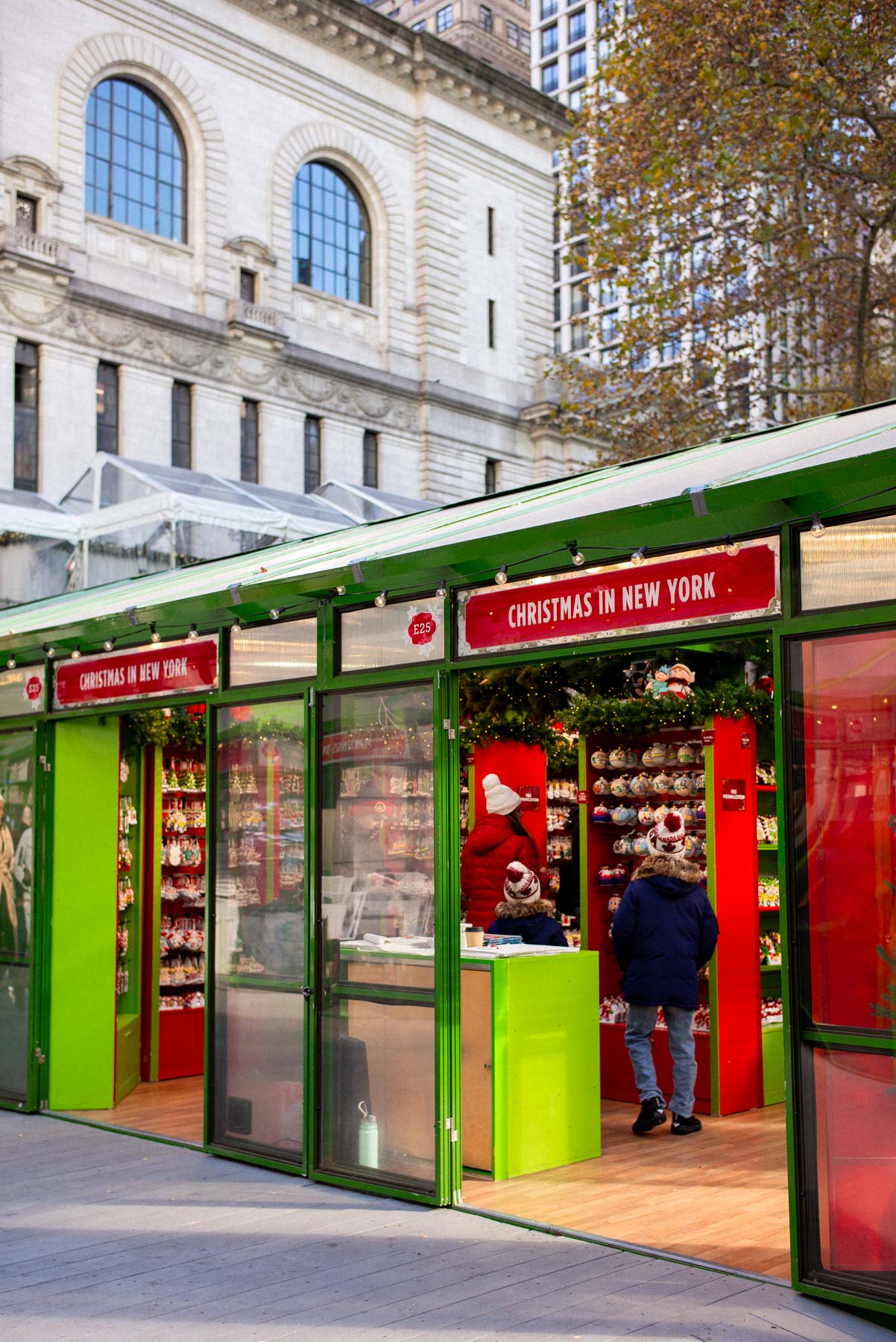 Two people browse a holiday market kiosk selling Christmas ornaments in front of the New York Public Library.