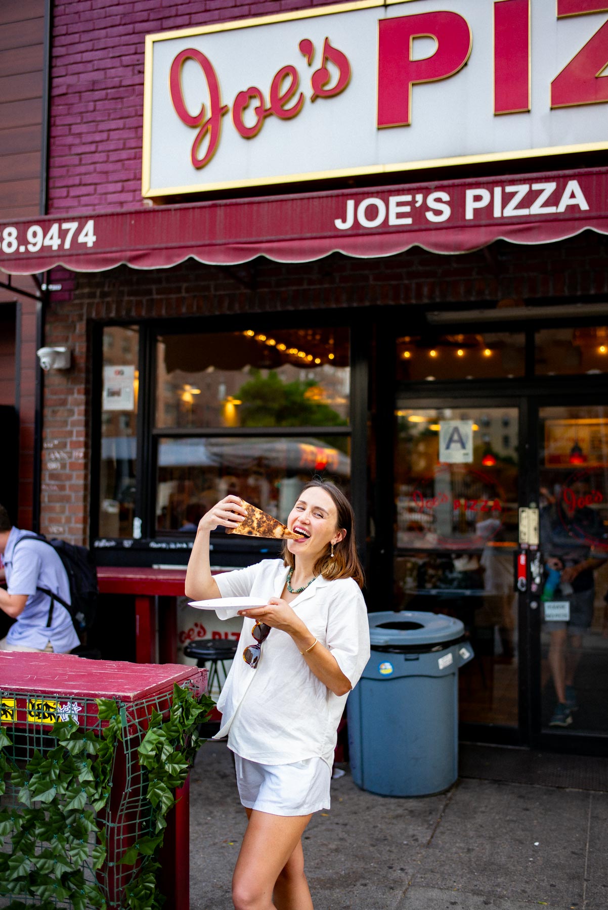 A woman eats a slice of pizza on a plate outside Joe's Pizza storefront.