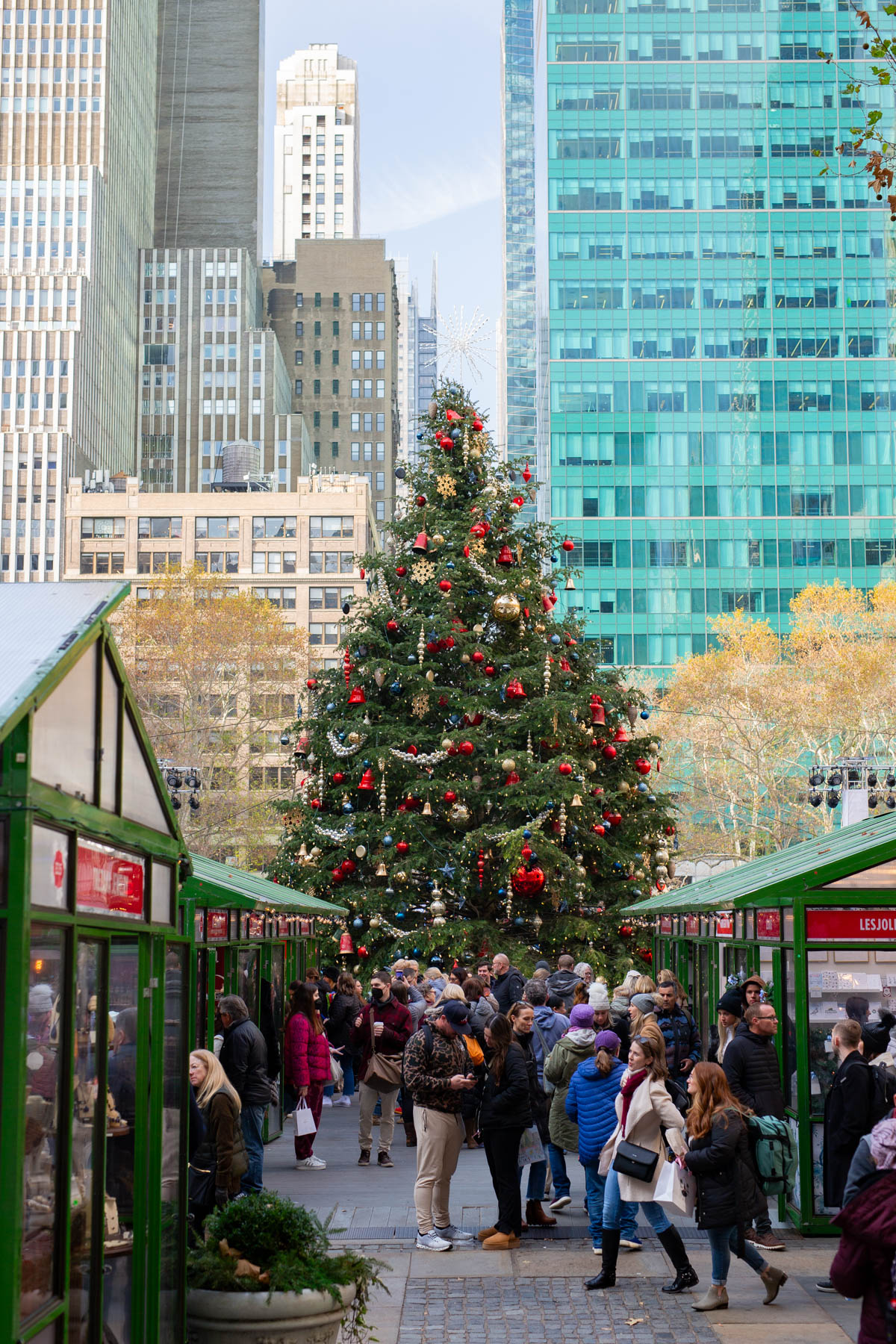 A decorated Christmas tree stands amid market stalls in Bryant Park with skyscrapers behind.