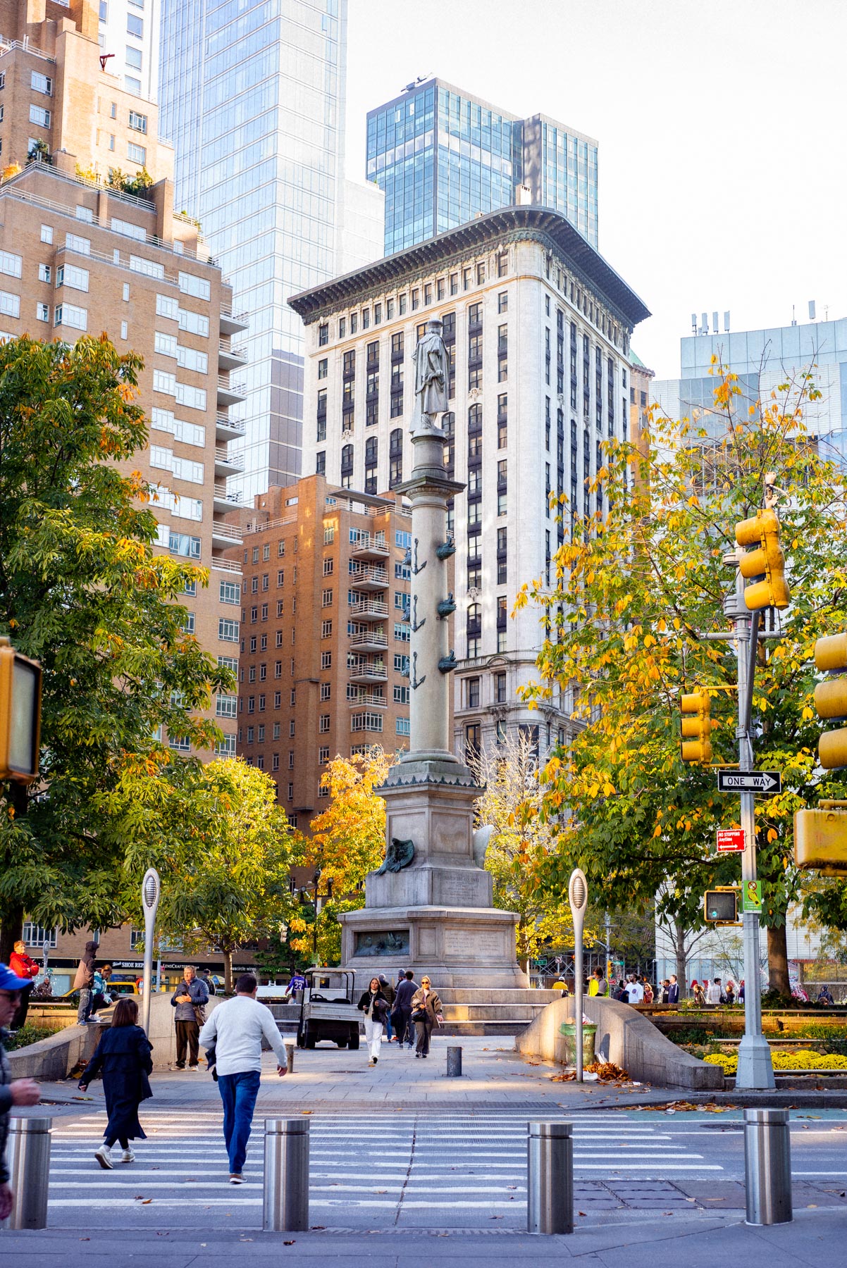 Columbus Circle monument stands amid fall foliage and surrounding buildings with pedestrians crossing the street.