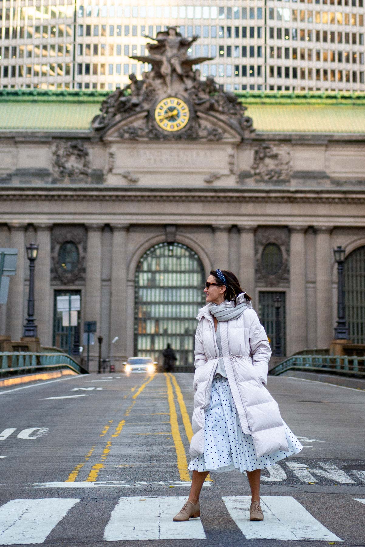 A woman in a puffer coat stands on a footbridge in front of Grand Central Terminal's clock and statue facade.